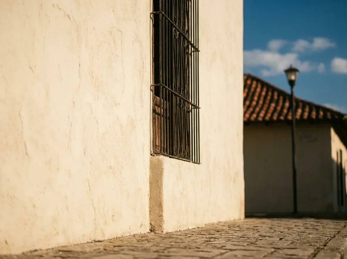 Esquina de una fachada encalada de color sólido con reja de hierro forjado sobre calle empedrada.